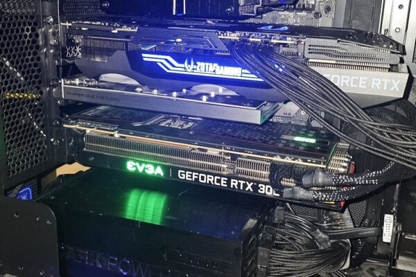 Blick in ein offenes schwarzes PC-Gehäuse von der Seite. Zwei Grafikkarten stecken übereinander in den PCIe-Steckplätzen: oben eine ZOTAC-Gaming-Karte mit blau leuchtendem Schriftzug, darunter eine EVGA GeForce RTX 3090 mit grün leuchtendem Logo. Zwischen beiden sitzt eine Erweiterungskarte für M.2-Speicher. Oben im Bild ein großer be-quiet-Luftkühler auf dem Prozessor, rechts geflochtene schwarze Stromkabel, unten ein Netzteil der Reihe Dark Power Pro. Die Karten liegen dicht beieinander, die obere verdeckt den Lüftereinlass der unteren fast vollständig.