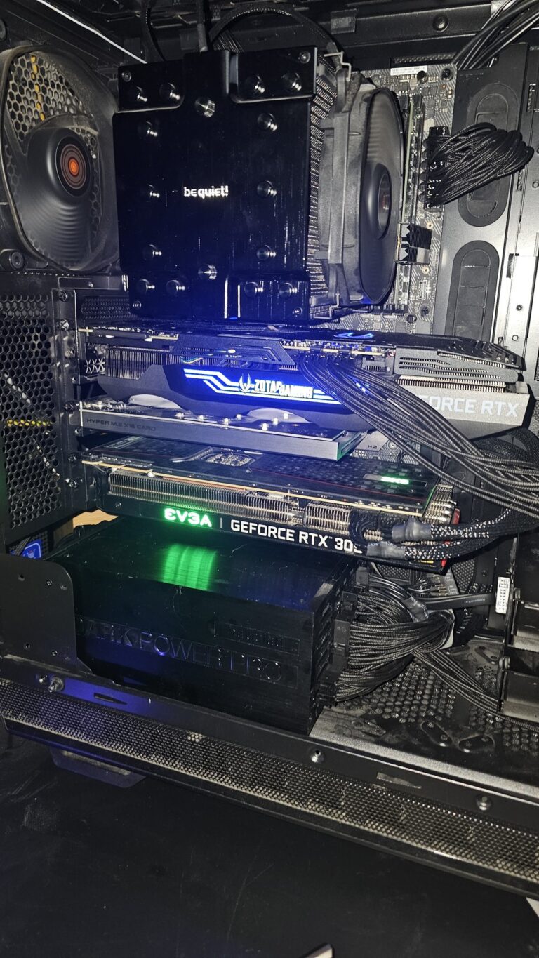 Blick in ein offenes schwarzes PC-Gehäuse von der Seite. Zwei Grafikkarten stecken übereinander in den PCIe-Steckplätzen: oben eine ZOTAC-Gaming-Karte mit blau leuchtendem Schriftzug, darunter eine EVGA GeForce RTX 3090 mit grün leuchtendem Logo. Zwischen beiden sitzt eine Erweiterungskarte für M.2-Speicher. Oben im Bild ein großer be-quiet-Luftkühler auf dem Prozessor, rechts geflochtene schwarze Stromkabel, unten ein Netzteil der Reihe Dark Power Pro. Die Karten liegen dicht beieinander, die obere verdeckt den Lüftereinlass der unteren fast vollständig.