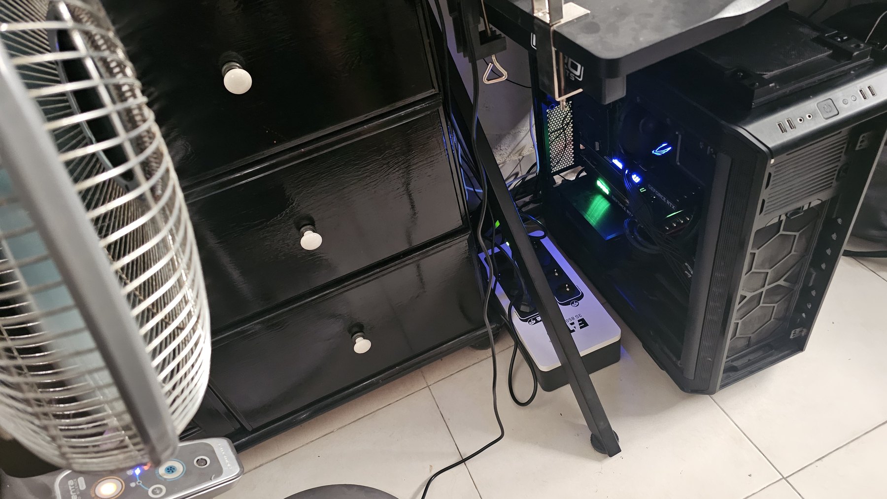 A large PC tower standing on a tiled floor next to a chest of drawers, with a pedestal fan pointed at it and a UPS on the floor